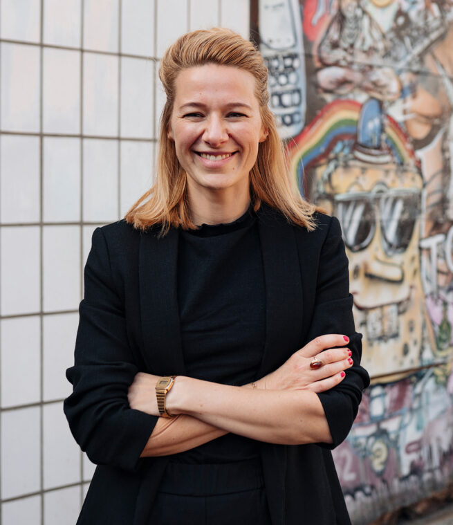 Britta Cornelißen, Blonde Frau in schwarzem Blazer mit überkreuzten Armen vor Graffiti Wand