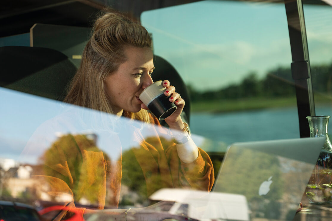 Frau aus Tasse trinkend im Van vor ihrem Laptop durch die Auto Scheibe fotografiert in der sich parkende Autos spiegeln
