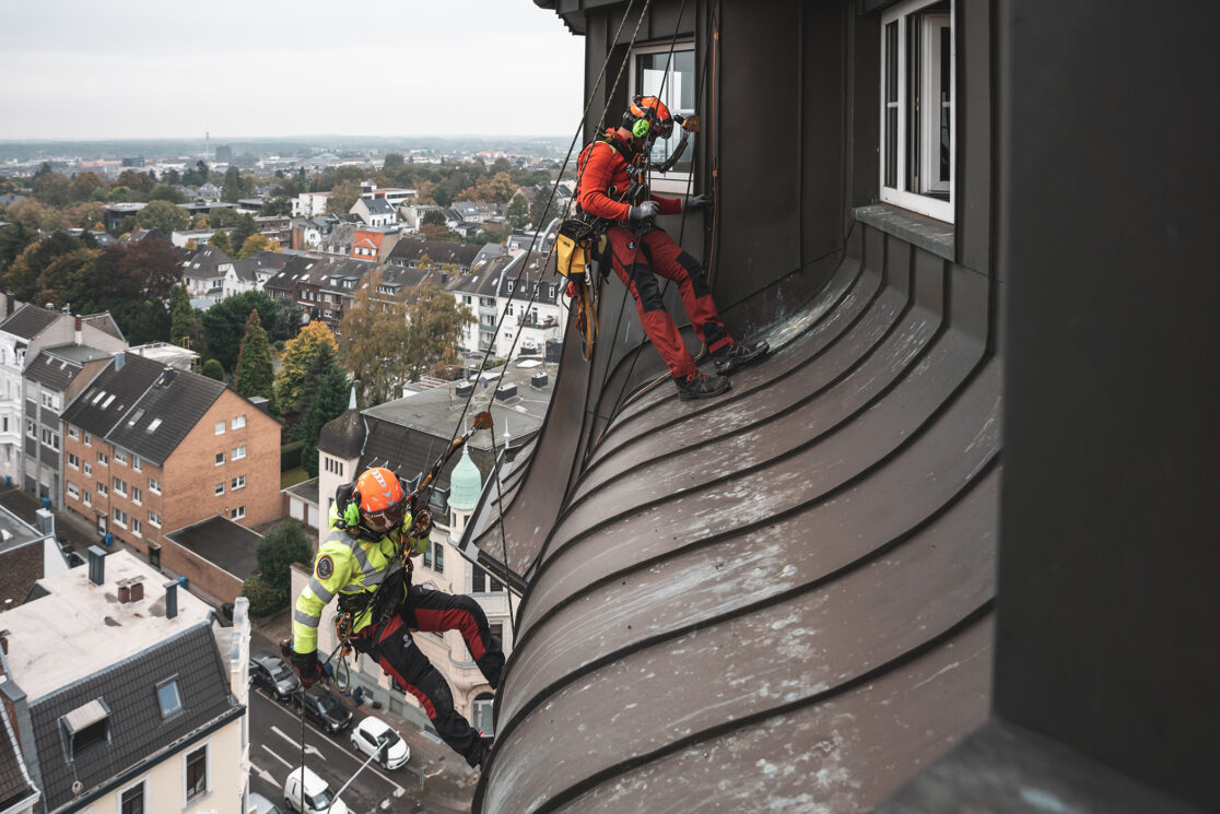 Zwei Industriekletterer in Sicherheitsausrüstung seilen sich vom denkmalgeschützten Wasserturm in Mönchengladbach ab. Stadt Mönchengladbach aus Vogelperspektive im linken Bildrand