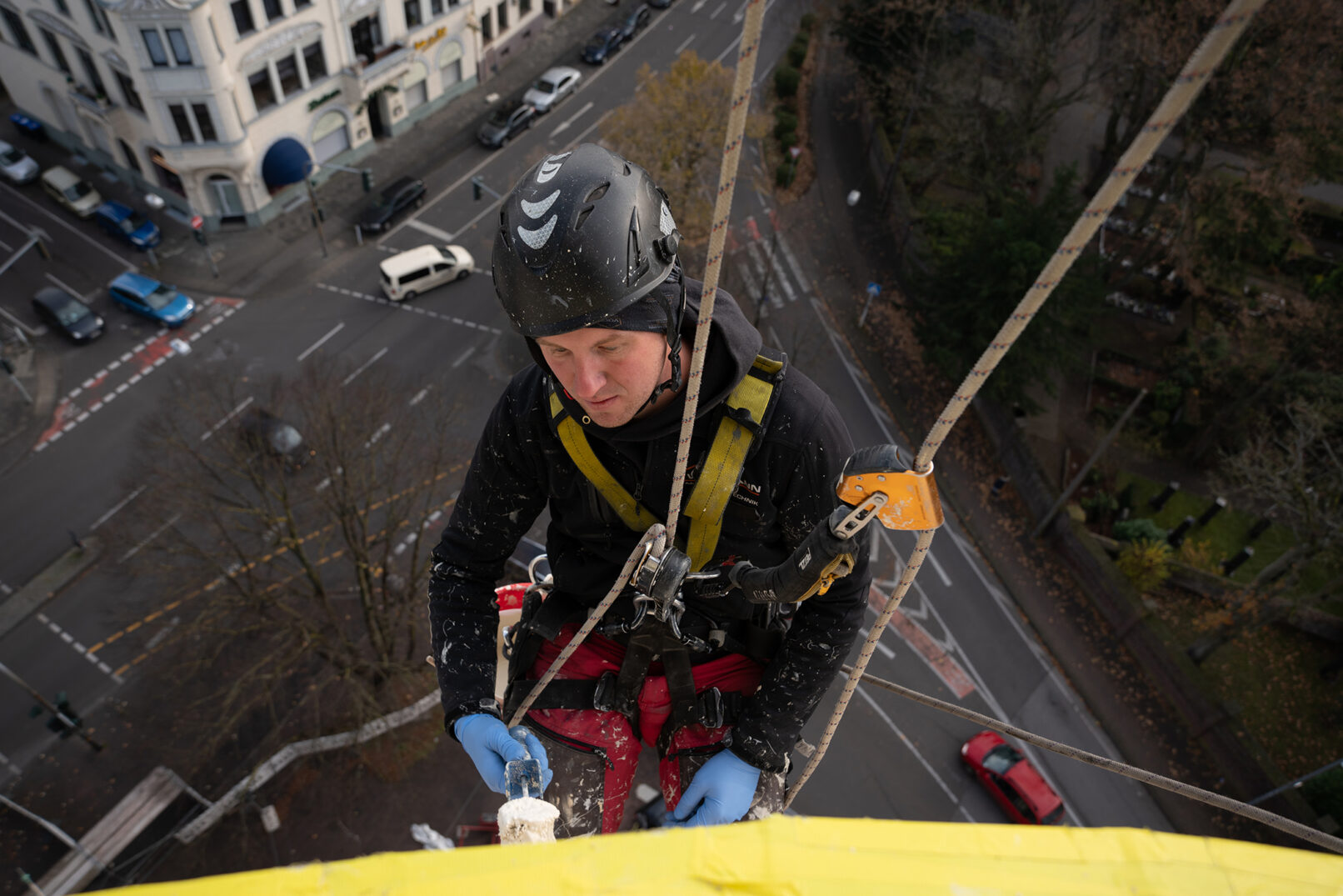 Fassadenkletterer im Klettergeschirr von oben aufgenommen mit Straße und kleinen Autos im Hintergrund