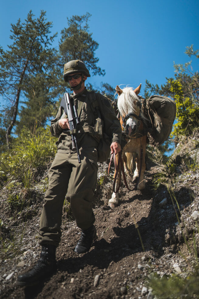 Von schräg vorne unten fotografierter Bewaffneter Soldaten des Bundesheeres Österreich, Tragtierstaffel marschiert einen steilen Pfad hinab und hält seinen hinter ihm gehenden Haflinger des Tragtierzentrums am Zügel.