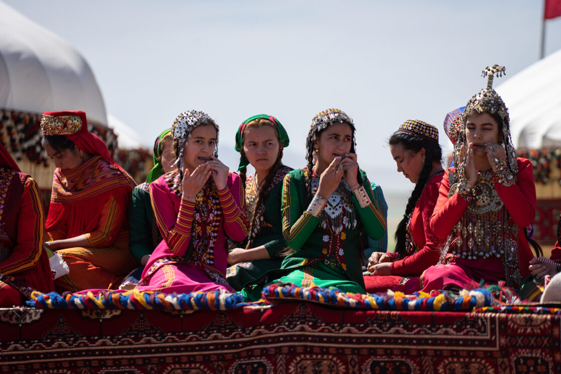 In bunter Landestracht gekleidete turkmenische Frauen sitzen auf einer mit bunten Teppichen angekleideten Empore und spielen Maultrommel