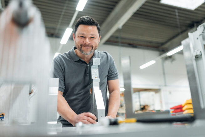 Businessreportage Rasch Verpackungsmaschinen Köln - Ein Mitarbeiter der Firma Rasch Verpackunsgmaschinen mit einem Bauteil in der Hand, in die Kamera lächelnd