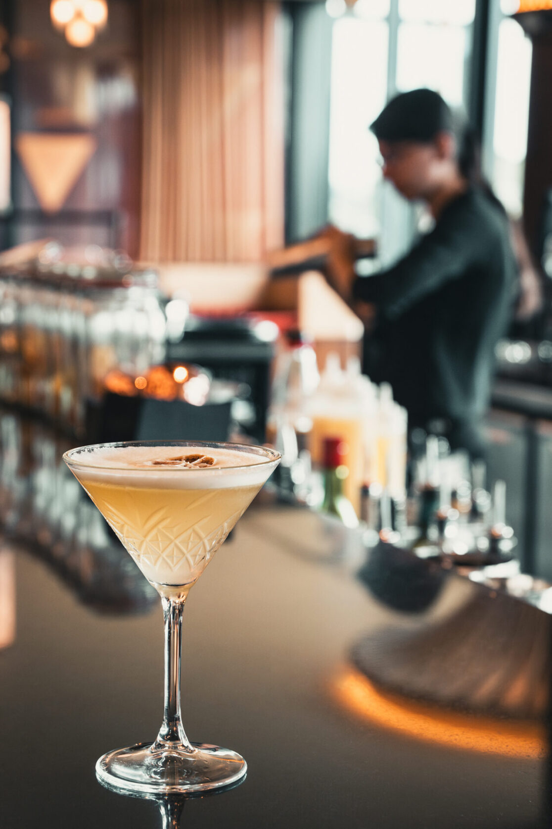 Cocktailglas auf einer Theke in der DOX Bar im Hyatt Regency Düsseldorf. Im Hintergrund ist die Barkeepern zu sehen, die einen Cocktail zubereitet.